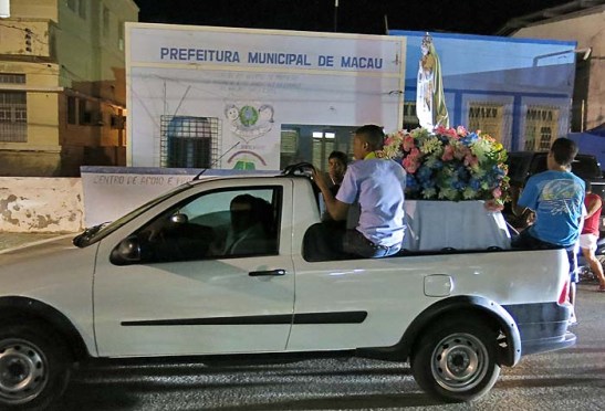A procissão com N.S. de Fátima em cima da camioneta e acompanhado por fiéis a pé e de carro pelas ruas da cidade. Na foto, o carro passa diante de uma unidade da Prefeitura de Macau.